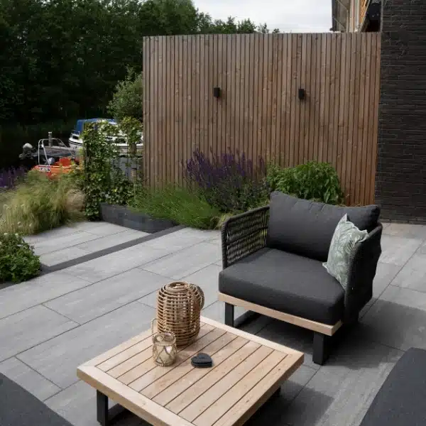 Moderne tuin met grijze terrastegels, houten loungeset en sfeervolle beplanting naast een houten schutting en uitzicht op een water met boten