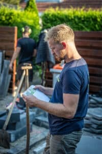 Tuinman bekijkt een tuinontwerp op papier tijdens werkzaamheden op een bestratingsproject, met gereedschap en materialen op de achtergrond.